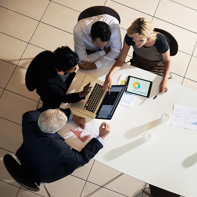 Four people sitting at a table looking at a laptop and tablet  with financial elements on them