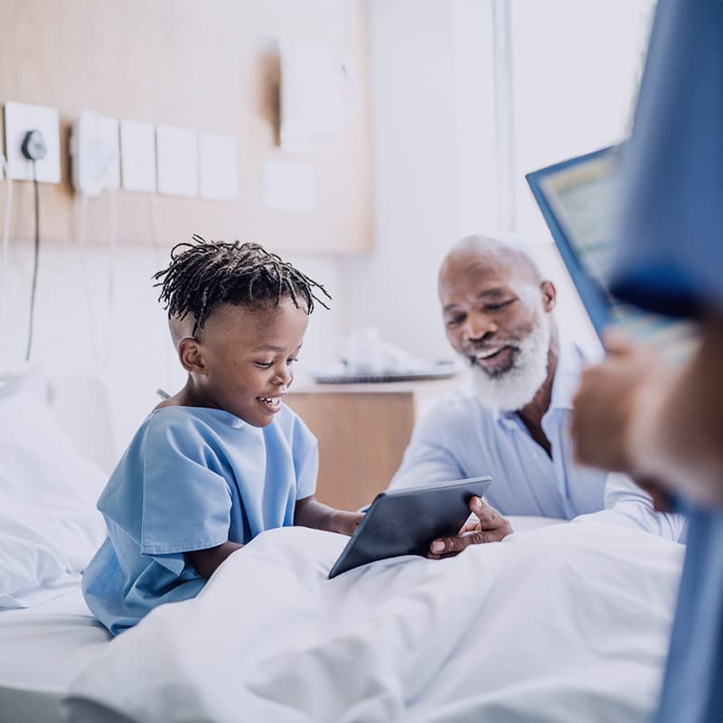 Father and son at a hospital bed with electronics: Golisano Childrens Hospital